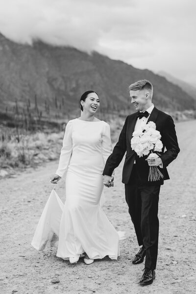 Natural photo of bride and groom walking along a dirt road holding hands with white roses and a moody mountain backdrop