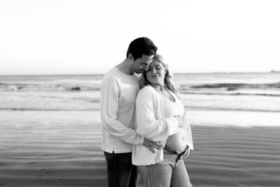 expecting couple standing close with eyes closed while holding mothers belly during a cozy beach maternity session in Orange County, California