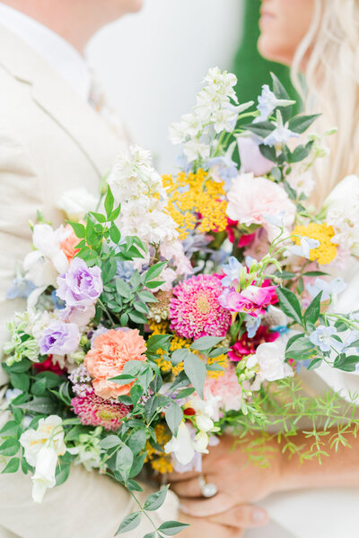 A vibrant, lush wedding bouquet featuring an organic mix of pastel and bold florals, including delicate blooms in pink, purple, yellow, and green.