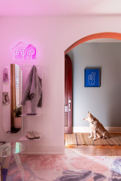A brightly lit neon sign hanging on the wall and a dog looking out an open front door.