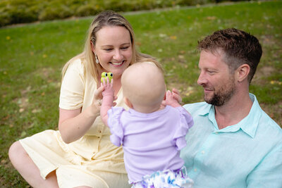 Playful family moment in Geelong park featuring parents engaging with their baby dressed in purple while sitting on the grass.
