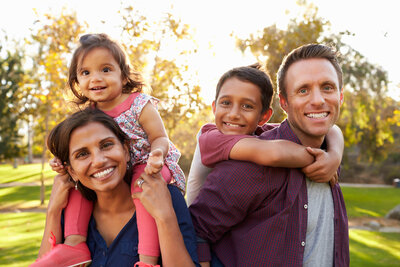 Two parents smile toward the camera, a sunlit natural scene in the background. Each parent has a child on their shoulders, also smiling at the camera.