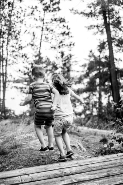 Two children walk hand in hand up log steps in a wooded clearing.