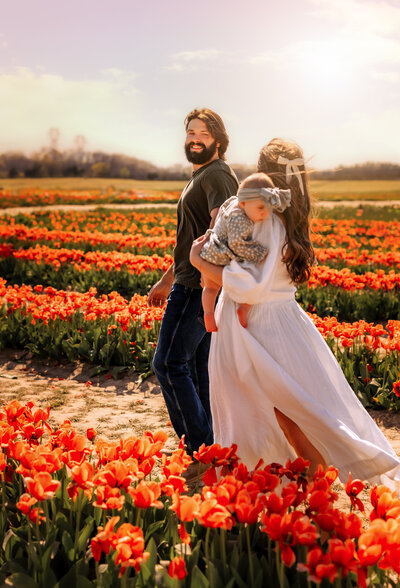 Mom, Dad, and two little girls are sitting on a blanket in a field with trees in a distance. They're all looking at one of the little girls smiling. 