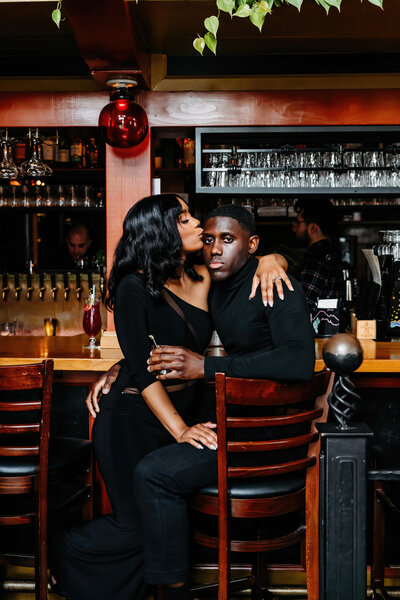 a BIPOC couple sitting at a bar, cuddled in each others arms
