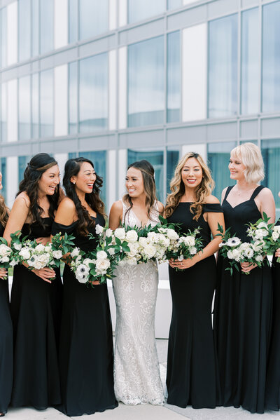 bride stands in the center surrounded by her bridesmaids wearing all black bridesmaid dresses holding their white and green bouquets from roots floral design