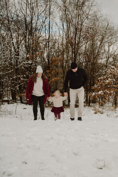 Mom wearing red, tan, and black walking with her husband who is in black and tan and young daughter wearing red and tan in the snow while smiling  in Severn Maryland.