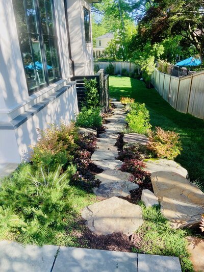 Beautifully landscaped side yard featuring a natural stone pathway, lush greenery, and vibrant plants leading towards a well-manicured lawn.