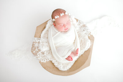 Newborn baby girl sleeping in a wooden basket. Wrapped in white swaddles, with bow headband.