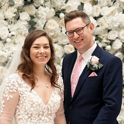 bride and groom standing in front of flower wall and smiling