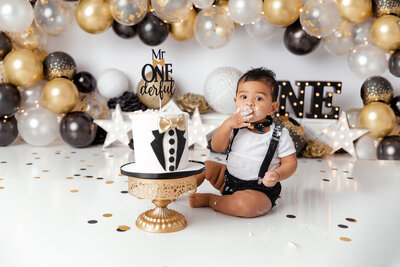 "Adorable baby celebrating his first birthday with a stylish Mr. Wonderful cake smash in a professional New Jersey photo studio. Captured by a skilled cake smash photographer, the scene features a golden and black balloon backdrop, a custom cake, and playful details for a memorable milestone session."