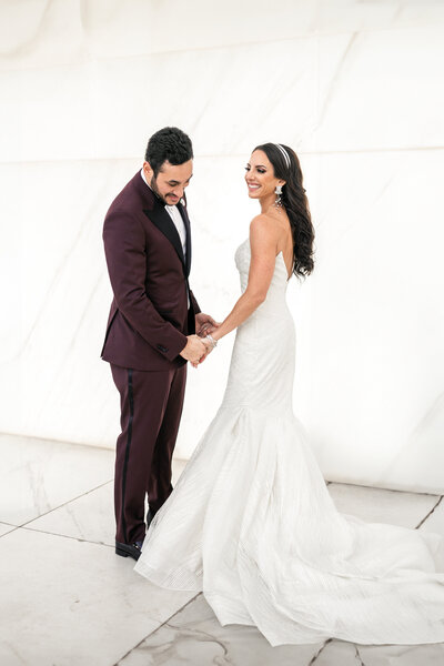 A couple stands indoors holding hands. The bride, in a strapless white gown with a flowing train, faces the groom, who is wearing a burgundy suit. They are smiling at each other, set against a light marble backdrop.
