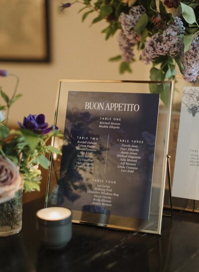 Close-up of a gold-framed seating chart reading "Buon Appetito," designed in a navy blue modern style for an elegant wedding at North & Navy, Ottawa. The chart complements the venue’s Italian-inspired charm, with florals and a candle adding warmth to the display. This stylish and functional seating arrangement sign blends personalization with a chic, old-world aesthetic, perfect for couples seeking unique, custom wedding stationery that enhances their event design.