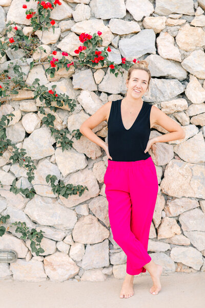 Caitlin Page Photographer Poses in Front of White Rock Wall with Red Flowers and Green Vines in a Black top and pink pants