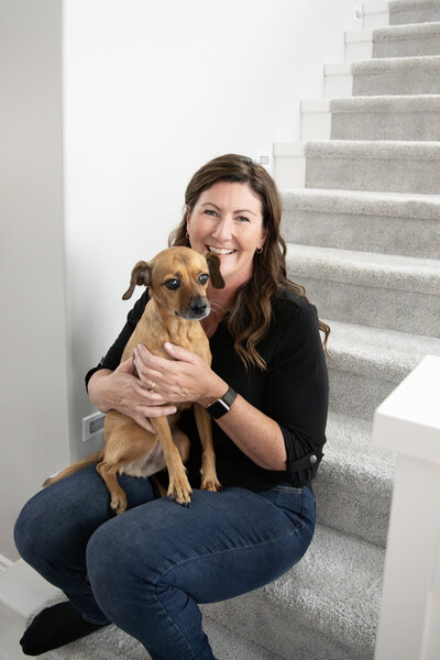 Michelle, founder of The Systems Café, sitting on a staircase and smiling while holding her puppy, Sadie, capturing a warm and personal moment that reflects her approachable and friendly nature.