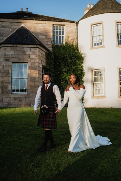 A wide photograph of a couple at golden hour. They walk down a hill with Logie Country House behind them.