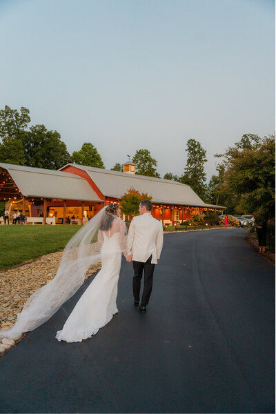 A bride and groom are walking towards the red barn at Hidden Hill for their wedding reception in Morganton NC.