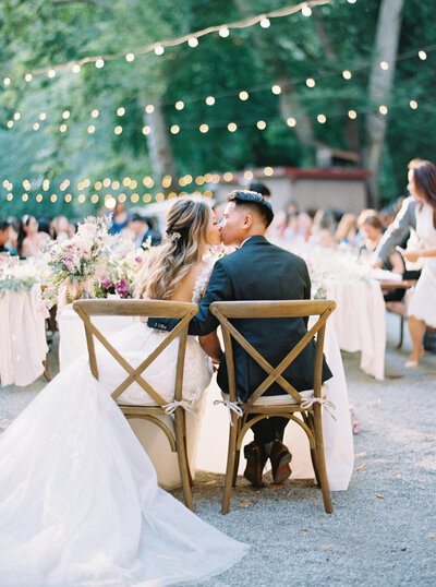 A couple at their wedding reception sitting at their sweetheart table kissing during sunset captured on medium format film.