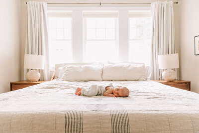 A newborn baby in pastel blue knit overalls lies on her tummy in the middle of her parents' king size bed in Somerville, MA