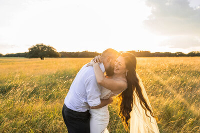 Groom kisses brides neck at sunset in meadow while she laughs.