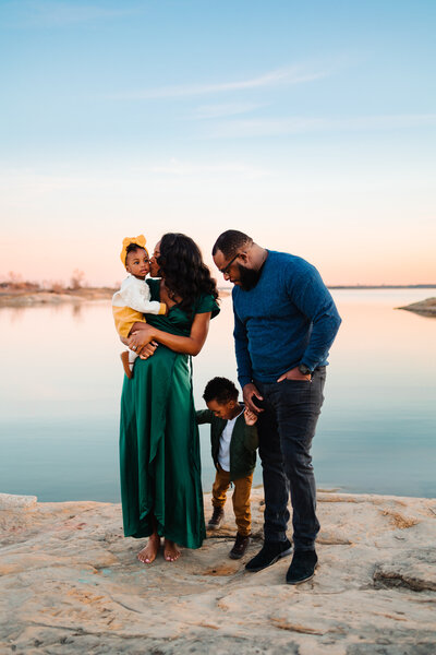 top-notch Albuquerque family photography with this captivating image of a family by a serene lake. The woman, dressed in a long green dress, holds a baby wearing a yellow cap and pants, who is gazing at the picturesque landscape.