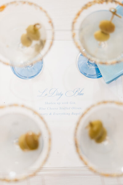 Crisp clean dirty martini glassis overlaying a blue and white wedding signature drink sign. 