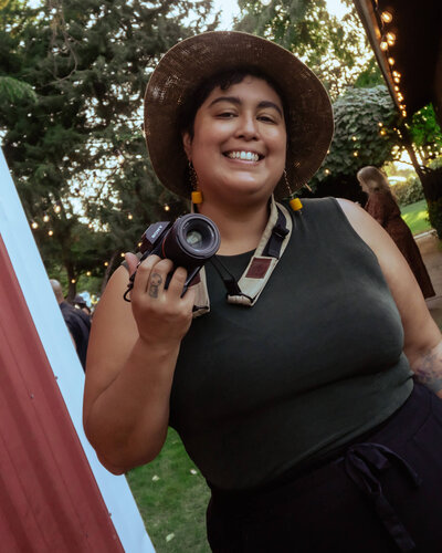 Cat Wheaton smiles at the camera while holding a camera. Representing their vendor profile for the Crush Collective wedding show.