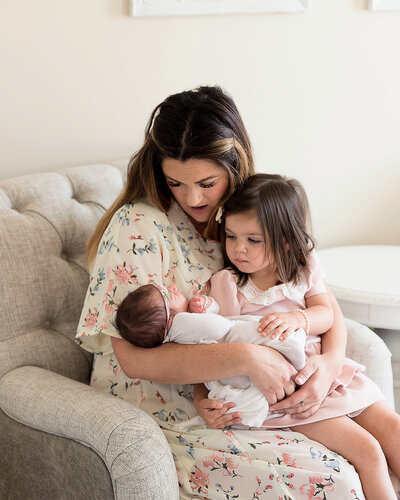 Lifestyle image of mother in a white floral gown holding her toddler and newborn