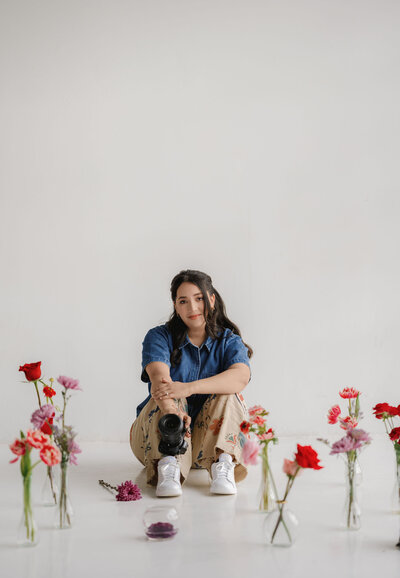 A photographer in a blue shirt and patterned pants sits on a white floor with a camera. She is surrounded by colorful flowers in vases, creating a serene, creative scene.