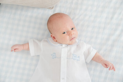Newborn baby in gown laying on a blue and white checkered crib sheet.