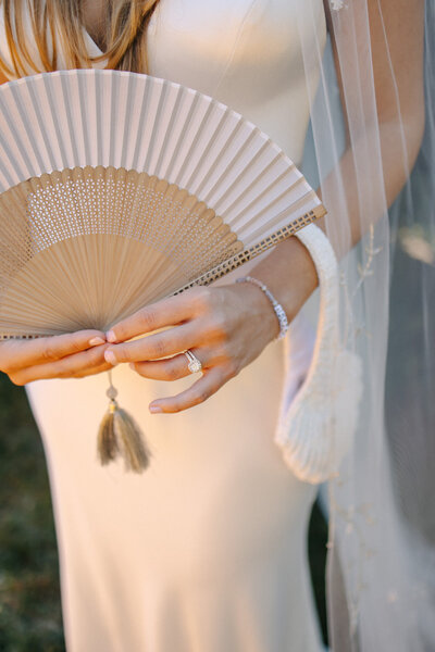 A close-up detail shot of a bride holding a light-colored pleated hand fan in front of her torso. She wears a sleek, strapless satin wedding dress with a delicate veil draping down her arm.