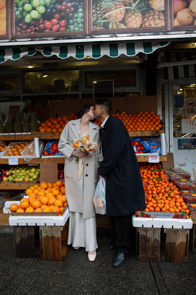 Brooklyn city hall elopement couple wearing kamperett kissing at corner fruit stand bodega in their neighborhood