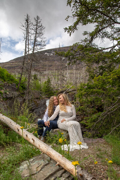 Two brides sit next to each other in a field of yellow flowers as their Montana elopement photographer take their photo.