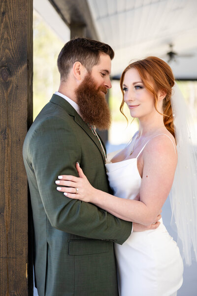 Bride and groom photograph.  Bride with large blue eyes and a white fitted wedding gown looking off in the distance towards the left.  Groom is lovingly looking at his Bride.  Groom holding bride at waist.  Bride holding on the grooms upper arms in an embrace