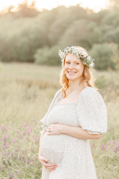 Pregnant Mama holding her belly in golden field at sunset. She is wearing a beautiful floral crown and is smiling a little off camera.