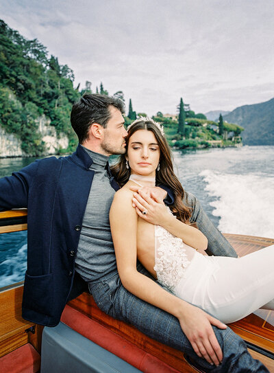 Engagement session on a sailboat. She is wearing a white halter top swimming suit sitting on the side of the boat with one leg down towards the water and the other one lifted straight out as she is kicking her legs and looking up at him. He is wearing white shorts with a white short sleeved dress shirt, standing behind her looking down at her while holding on the the rope of the sail. You can see water and a bridge behind them. Photographed by wedding photographers in Charleston  Amy Mulder Photography