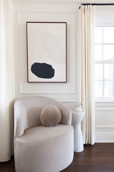 A quiet, neutral-colored reading nook featuring abstract art,  marble side table, and curved chair.