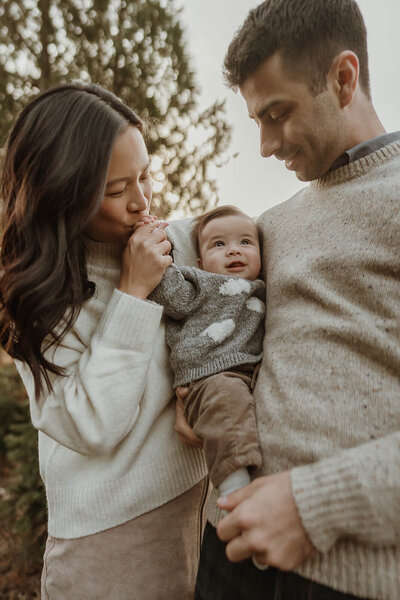 Orange County fall family photography of parents holding their baby outdoors, mother kissing baby’s hand, all wearing cozy neutral sweaters during golden hour.
