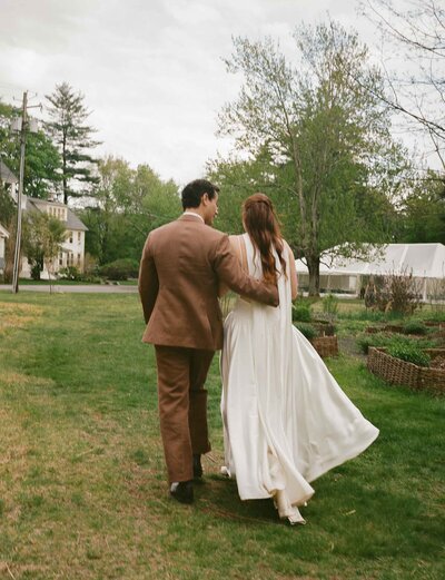 A bride and groom stroll hand in hand through a lush green field, surrounded by nature on their wedding day.