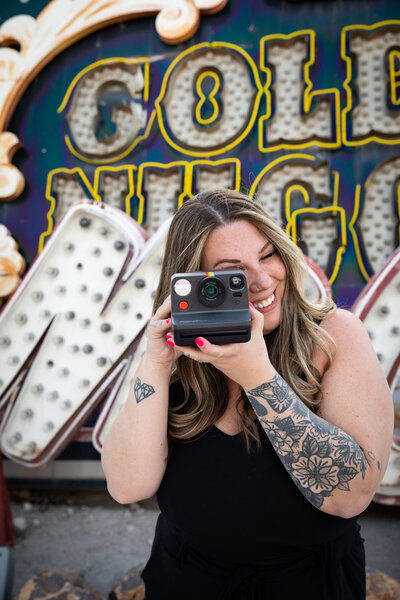 A woman with tattoos capturing a photo in front of a vibrant neon sign at an Austin photo studio.