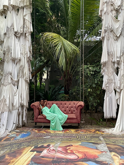 Nadia Forte lounges on a vintage sofa in a lush, tropical setting, surrounded by artistic drapery and intricate decor. As the founder of Nadia Forte Travel & Events, she specializes in bespoke luxury travel, destination event planning, and exclusive high-touch experiences worldwide