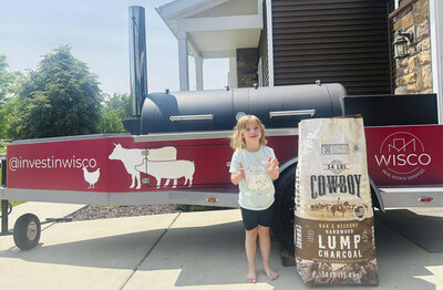 Girl next to brisket smoker trailer and bag of lump charcoal