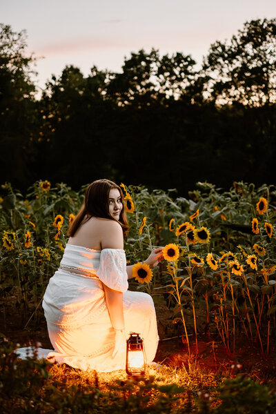 Senior teen girl looking over shoulder in sunflower field wearing white dress as sun is setting with lantern next to her in Maryland.