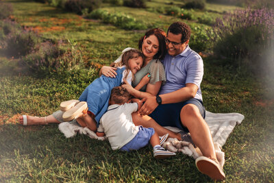 A mom is standing in the middle smiling while looking down while two little girls are running around her with their hair blowing in the wind while wearing flowy dresses. Theres leaves on the ground and water in the background and a tree branch above them.
