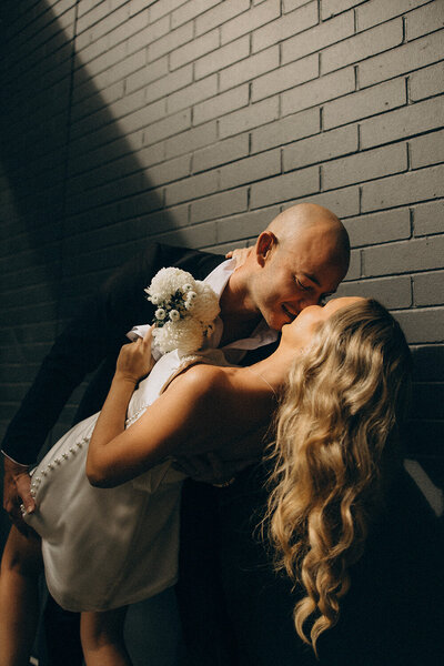 Fun, romantic Sunshine Coast elopement photo of groom dipping his bride beside a brick wall.