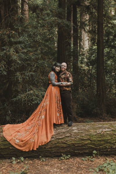 queer wedding couple, one in an orange wedding dress, standing on a fallen tree edited in a dark and moody style