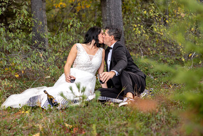Bride and Groom sitting in the forest on a blanket with champagne and toasting.