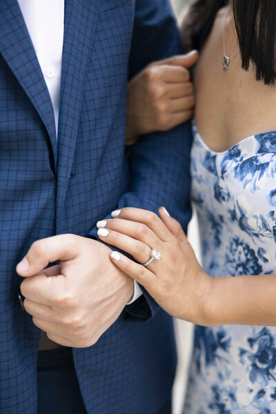 A couple embracing, with the woman wearing a blue floral dress and showing a square-cut engagement ring. The man is in a blue checked suit. Her hands gently rest on his arm, which is crossed in front of him.