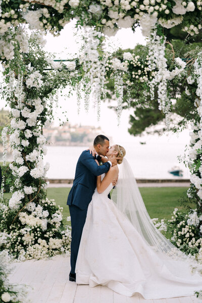 Newlyweds share a kiss under a lush white floral arch by the water, surrounded by greenery and sunshine