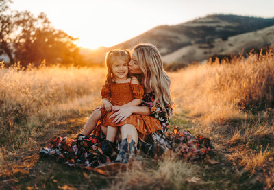 Family Photographer, a mother kisses her daughter as she sits on her lap in a meadow